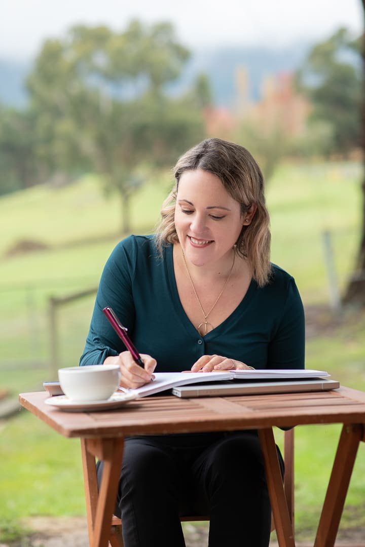 A candid personal branding portrait in which a Woman sits in an outdoor setting and looks down at a pen and paper. She is writing at a coffee table.