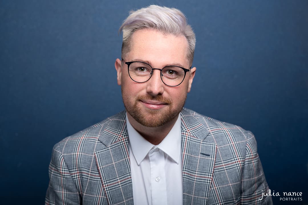 Man on navy background wears a suit and glasses, for a relaxed yet serious corporate headshot