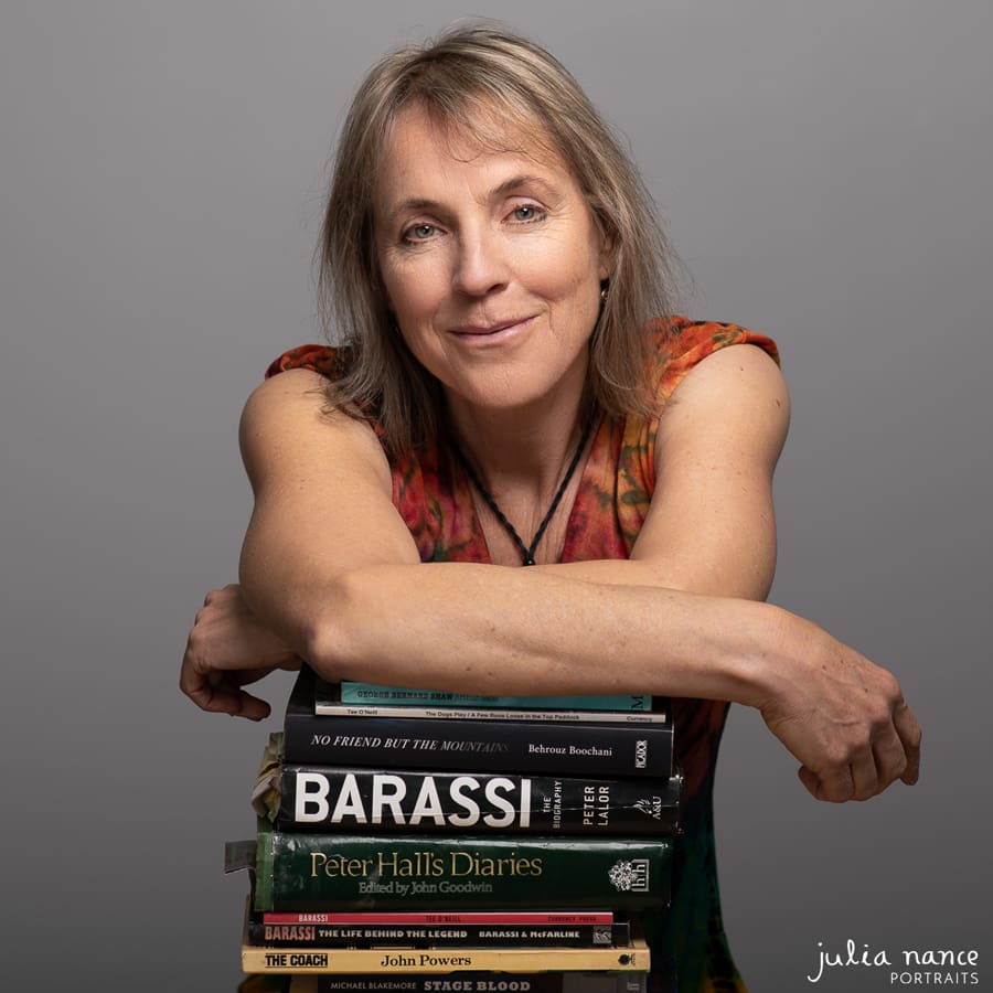 Author portrait of woman who has her elbows leaning on a pile of books, taken in a studio environment.
