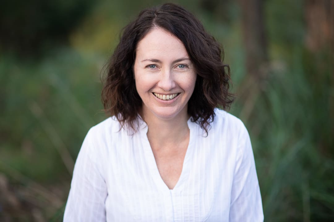 Woman outdoors smiles at the camera for a relaxed headshot. She wears a white top and has brown shoulder length hair.