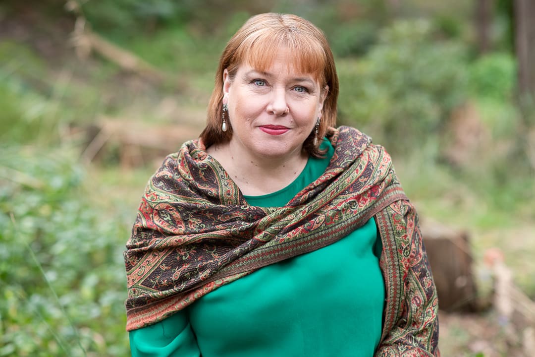 A relaxed outdoor headshot of a woman sitting down. She wears a green top with a scarf around her shoulders.