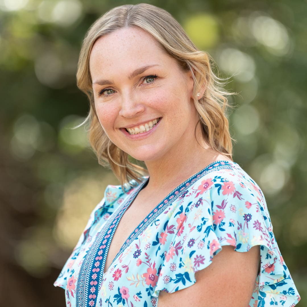 Woman poses for corporate headshot in an outdoor setting. She wears blue ruffled sleeves and behind her the background is soft and blurred.