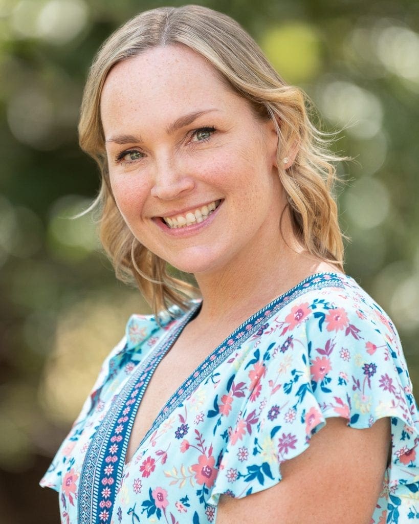 Woman poses for corporate headshot in an outdoor setting. She wears blue ruffled sleeves and behind her the background is soft and blurred.