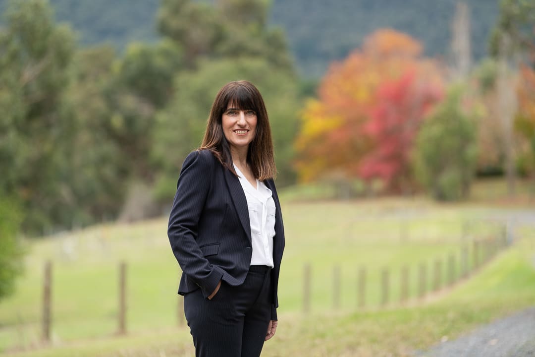 Woman in outdoor setting has hands in her pockets for a calm branding portrait. There are autumn trees behind her.