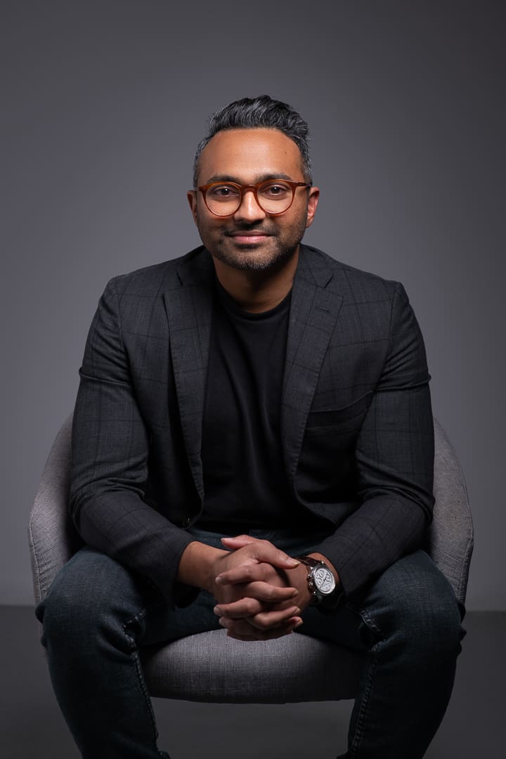 Contemporary corporate branding portraits of man sitting down. He has his hands clasped and a gentle expression on his face. The image has dramatic lighting, and the man wears a professional blazer and suit pants.