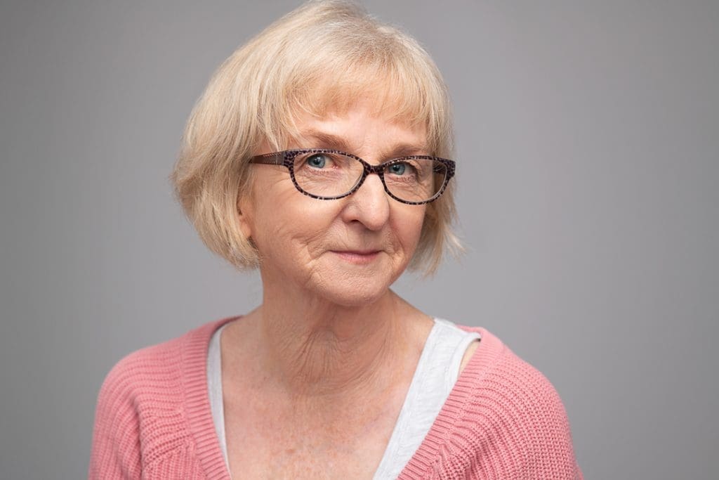 Actor headshot of a Woman photographed in a studio wears a pink cardigan and glasses. She has a knowing look in her eye.