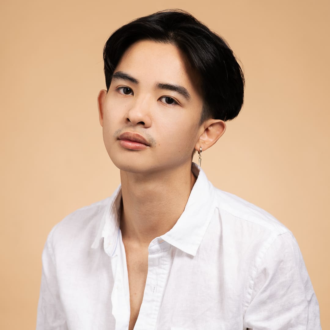 Actor headshot of young man wearing a white collared shirt. The background is a peach apricot, and he has a serious expression.