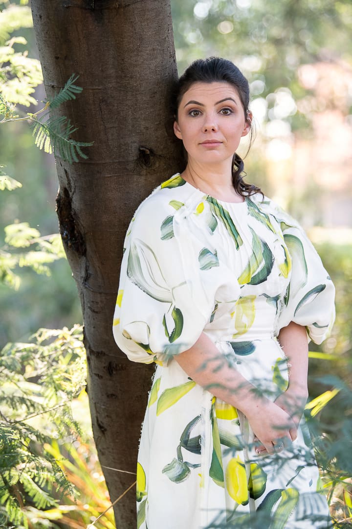 A personal branding portrait of a woman leans against a tree outside in a natural outdoor setting in melbourne and smiles to camera.