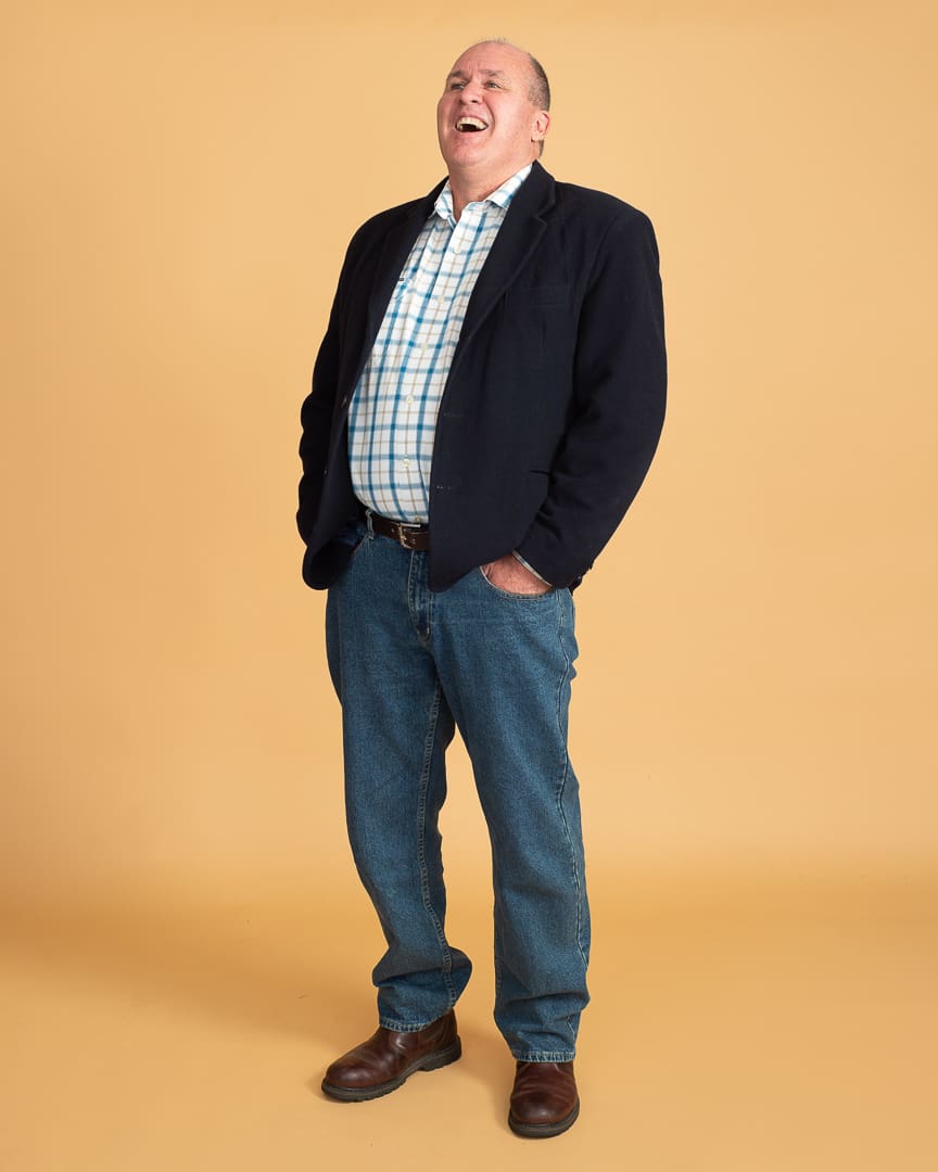 A personal branding portrait of melbourne man who laughs off-camera with his hands in his pockets. The portrait is full length, and is taken on a seamless orange background. He wears jeans, a checked shirt and a navy jacket.