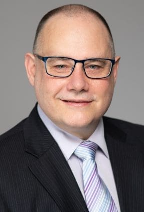 Professional corporate headshot of melbourne accountant wearing glasses and a formal suit. He smiles to camera on a light grey studio background.