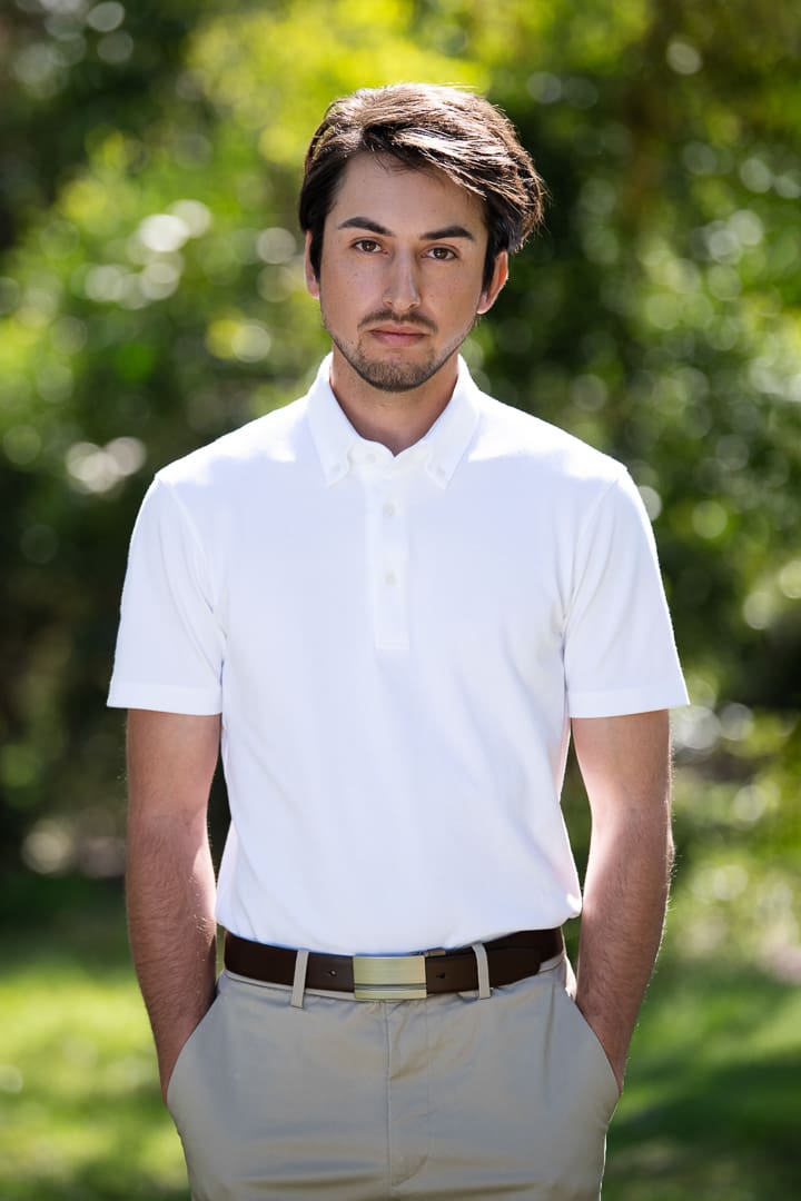 Melbourne model portfolio portrait of young man in white polo shirt, taken outside. He has a serious expression and the sun shines from behind him.