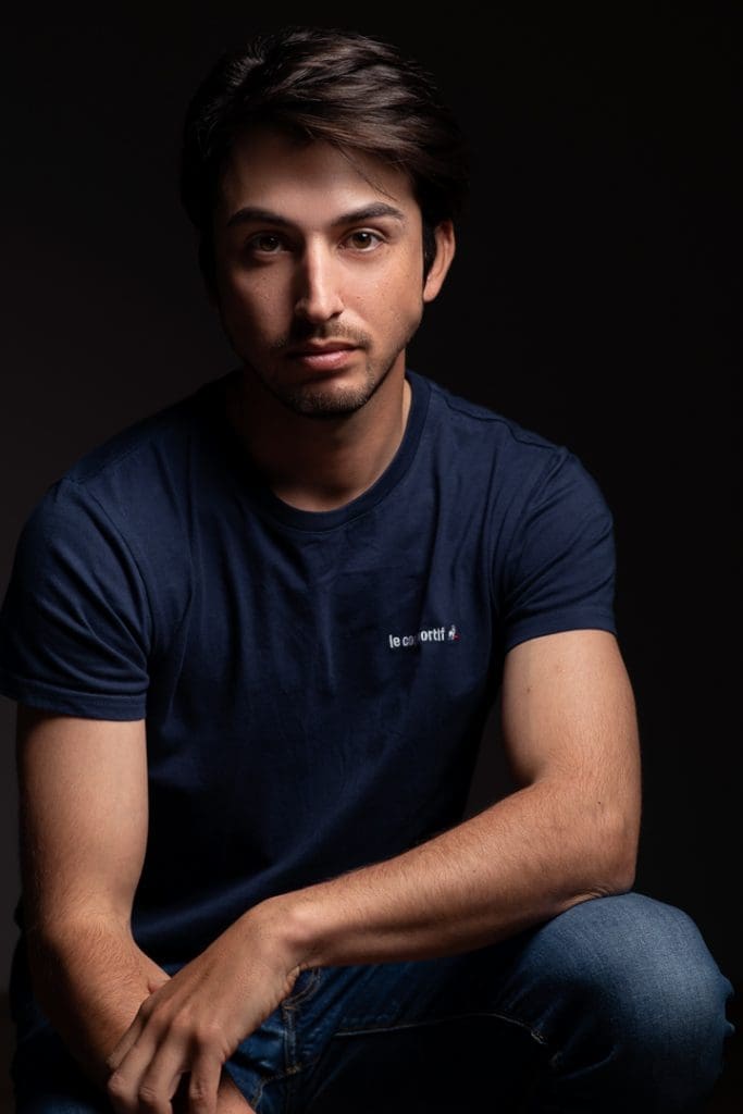 Melbourne model portrait taken on a dark studio background with edgy lighting. he looks directly to camera while leaning on his knees in a seated position.