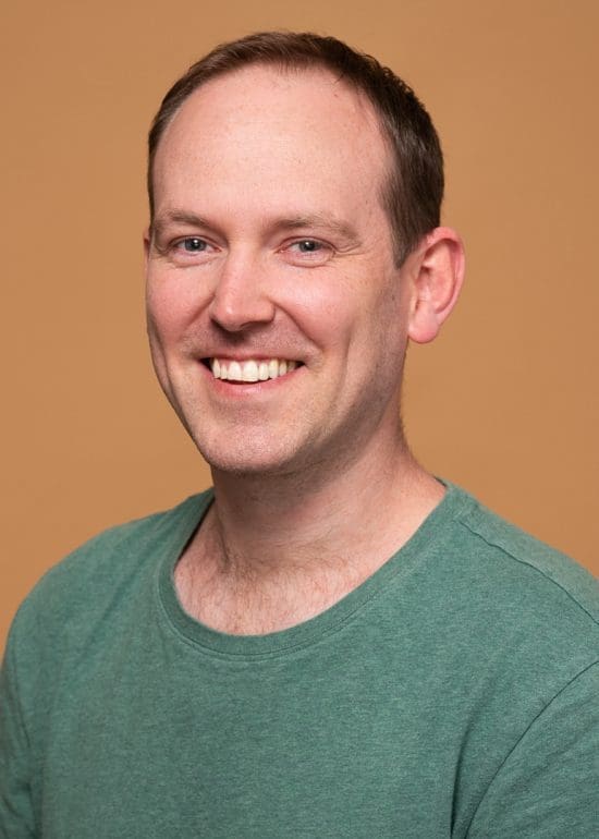A relaxed linkedin headshot of melbourne man wearing a green tshirt with an orange background behind him. He has a natural smile and looks at the camera.