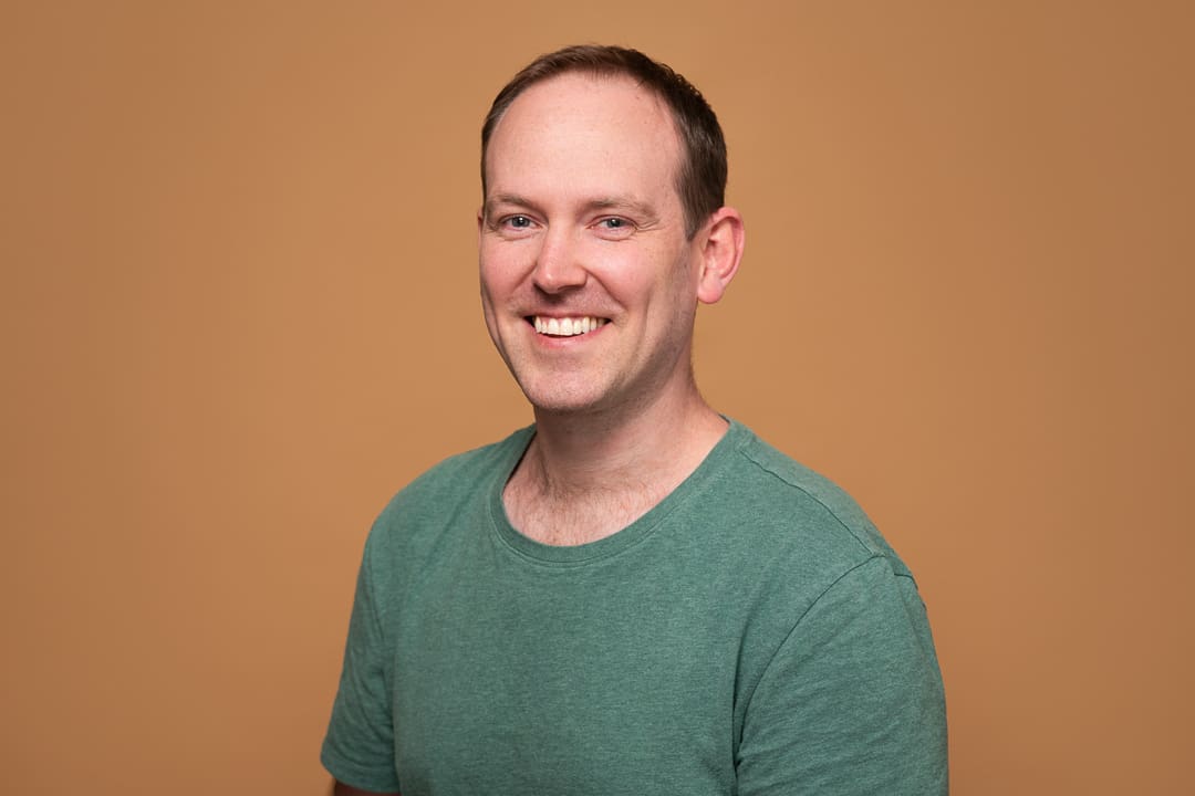 A relaxed headshot of peter, wearing a green tshirt on an orange background. he smiles to camera in my melbourne studio.