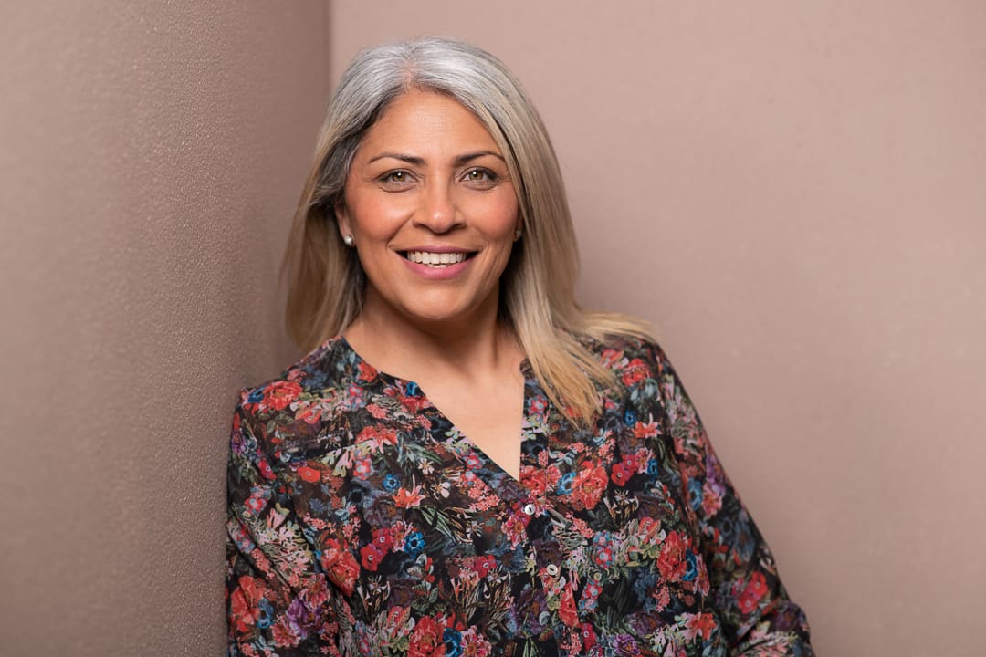 Relaxed headshot of woman leaning against an earthy wall. She smiles to camera wearing a floral blouse.