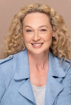 Melbourne actress smiling for a relaxed actor headshot. She wears a blue blazer and is in front of an earthy brown background in a studio.