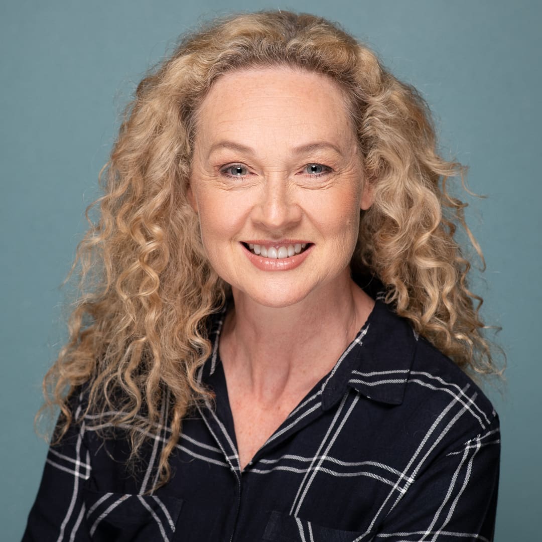 Smiling actor headshot of woman with blond curly hair taken in a studio on a blue background.