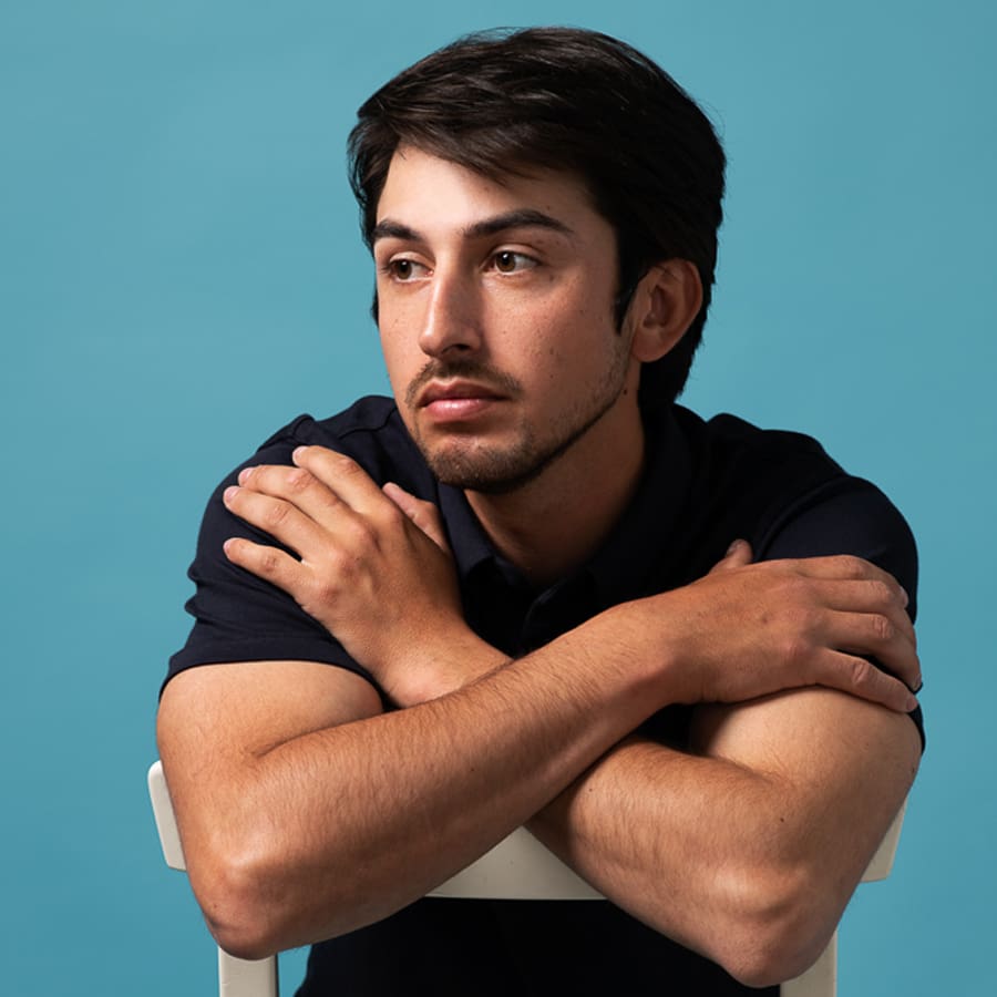 Model portrait of man leaning over the back of a chair on a blue background. He is looking away from the camera.