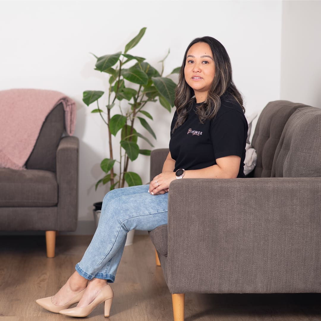 A personal branding portrait of melbourne mortgage broker seated on a couch with a green plant behind her. She wears a branded tshirt, jeans and heels.