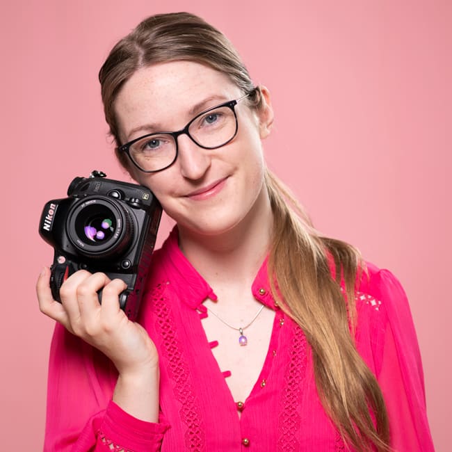 Professional headshot of Julia nance holding a camera next to her cheek, smiling to camera on a pink background, wearing pink.