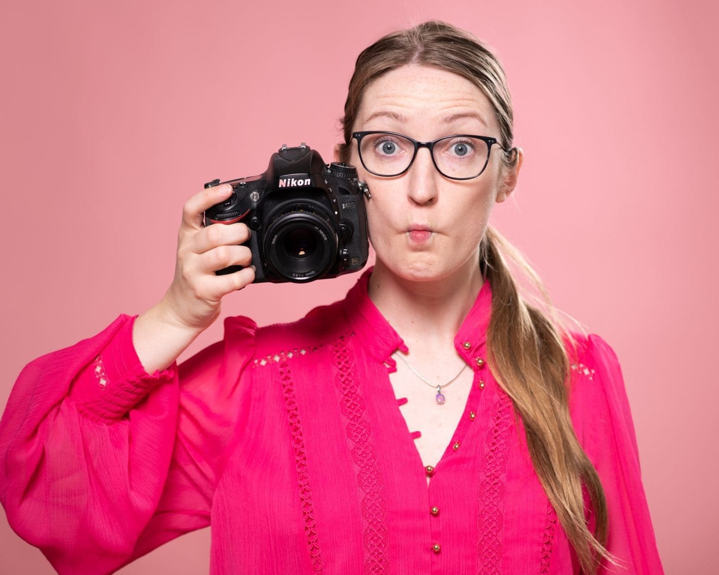 Portrait of Julia Nance on a pink background, wearing a pink shirt and holding a camera with a quirky expression (fish face).