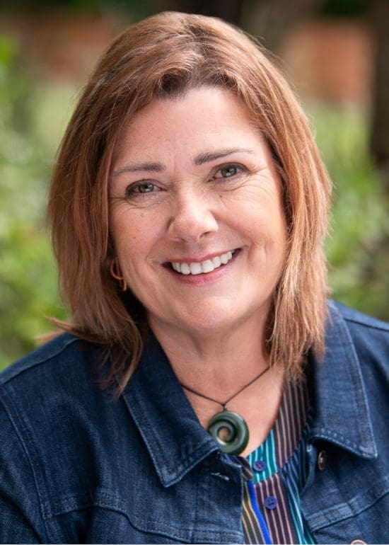 Relaxed linkedin headshot of woman smiling to camera in denim jacket. The photograph is taken in natural light with an outdoor setting of greenery.