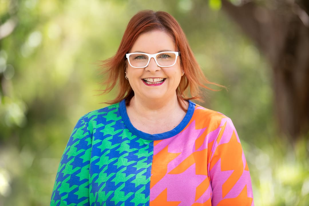 Personal branding portrait of woman with red hair and white glasses, smiling at camera. Her headshot is colourful and bold, captured in natural light outdoors.