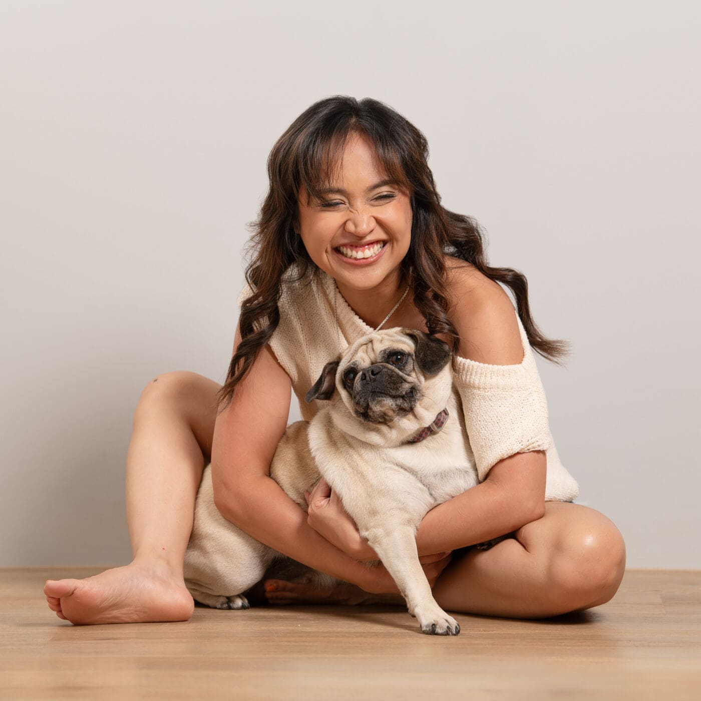 Woman laughs off camera while sitting on the floor, she holds her small pug in her lap.