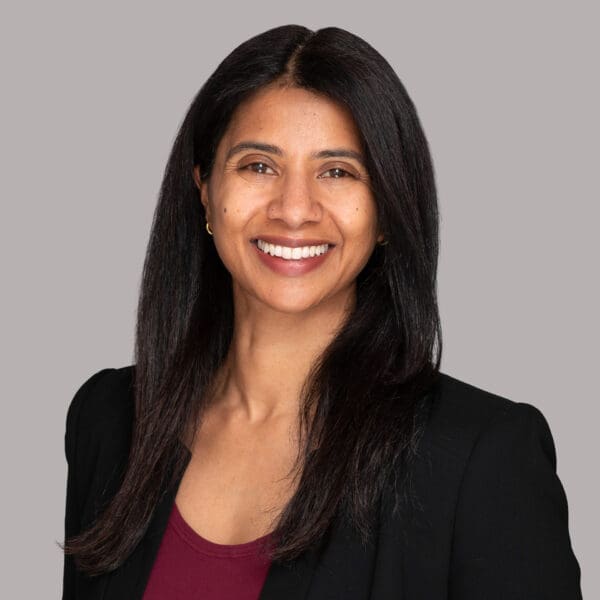 Woman smiles to camera in corporate portrait. She wears a blazer and is photographed on a light grey background.