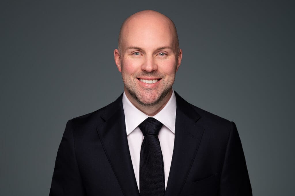 Corporate headshot of a man in a suit and tie. He is facing front-on and smiles to camera on a blue-grey background.