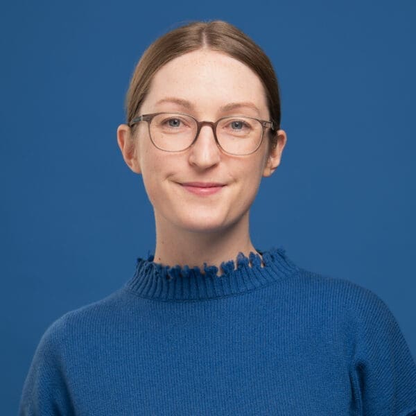 Portrait of Julia Nance wearing a royal blue jumper on a matching blue background. She smiles to camera.