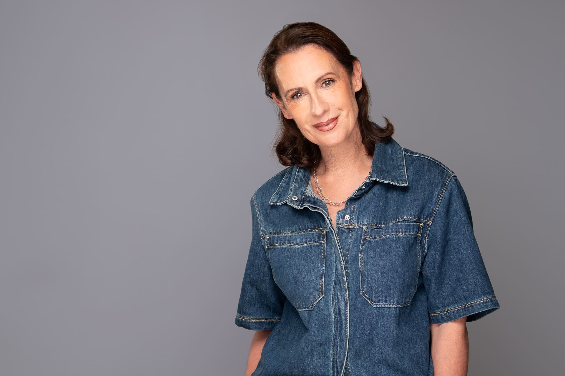 Woman smiles to camera with tilted head. She wears a denim top and the background is a cool grey/blue.