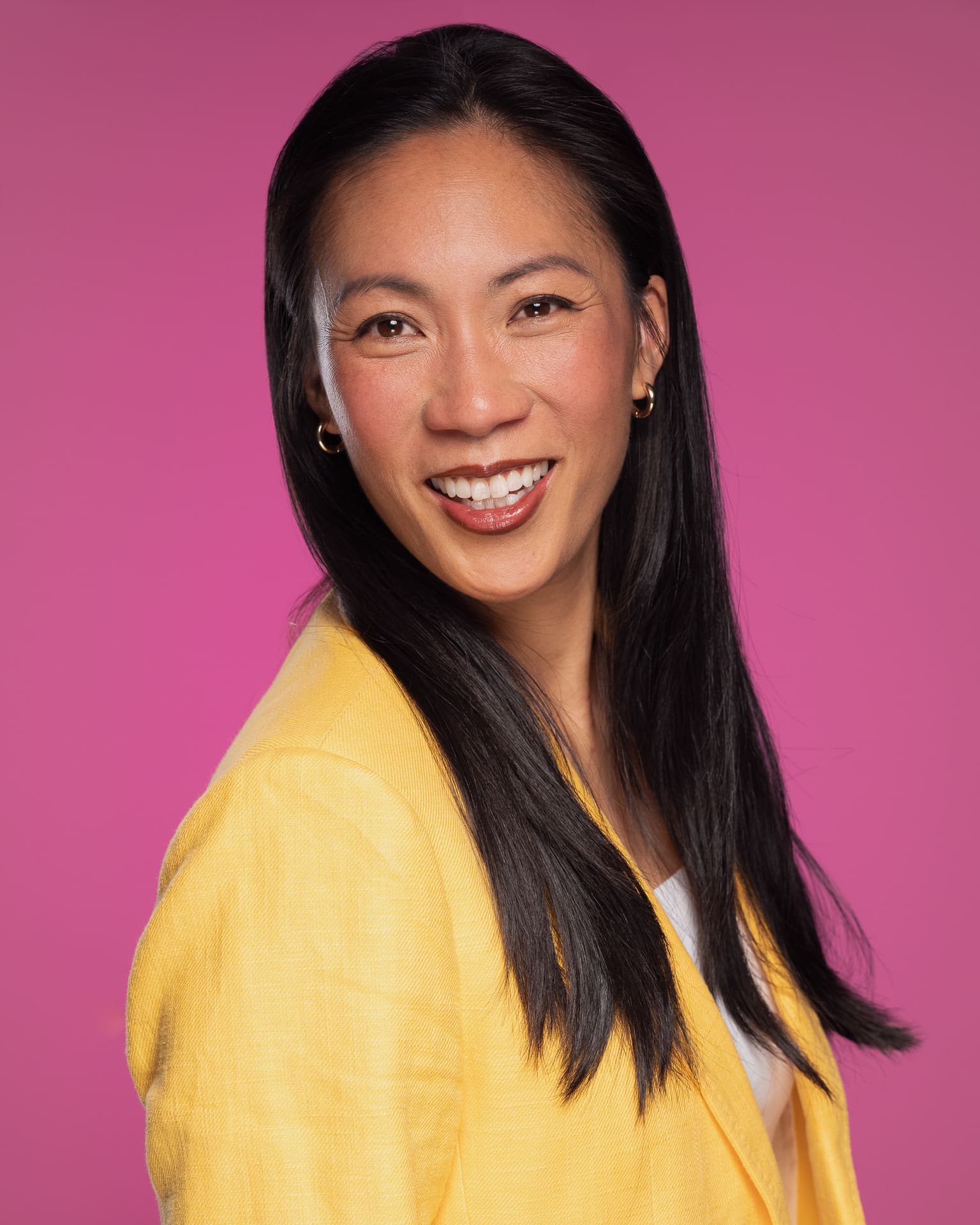 Woman smiles to camera wearing a yellow blazer on a pink background.