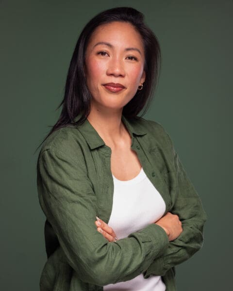 Woman looking to camera with arms crossed and slight smile. She wears green and is photographed on a green background.