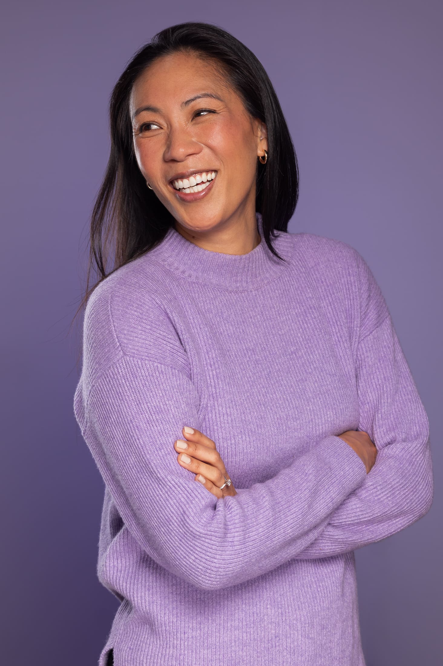 Woman wears lilac jumper on a purple background, with her arms crossed and smiling away from the camera