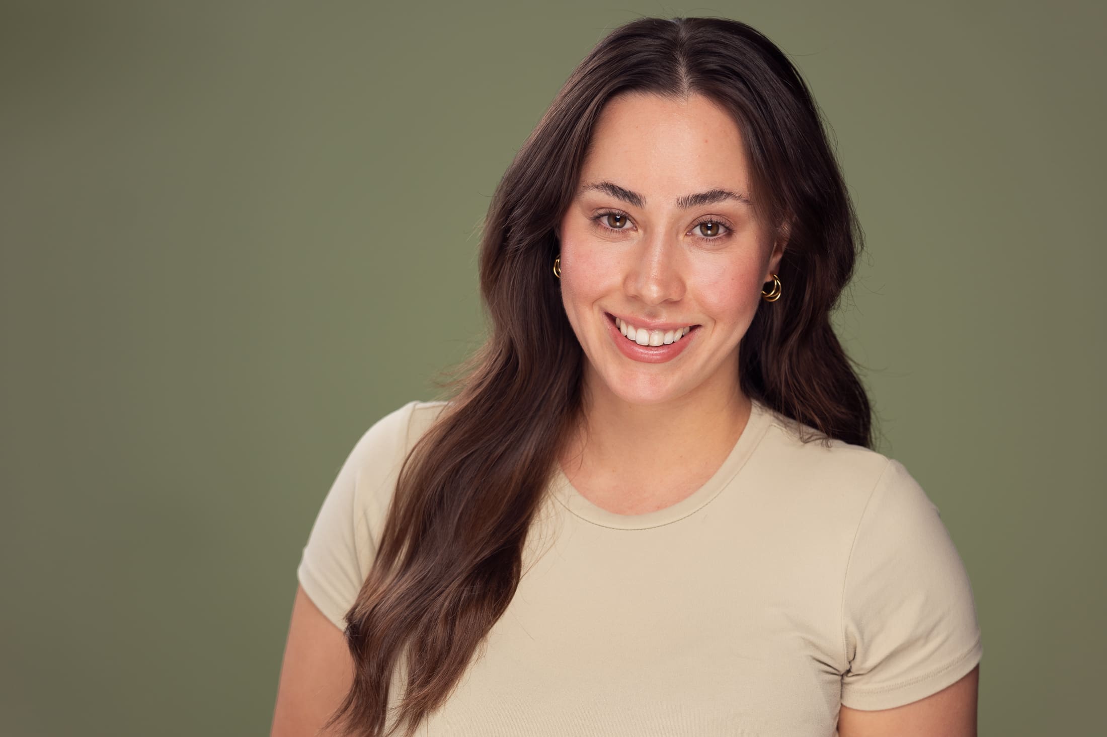 Woman smiles to camera, wearing a light green tshirt photographed on a mossy green background.