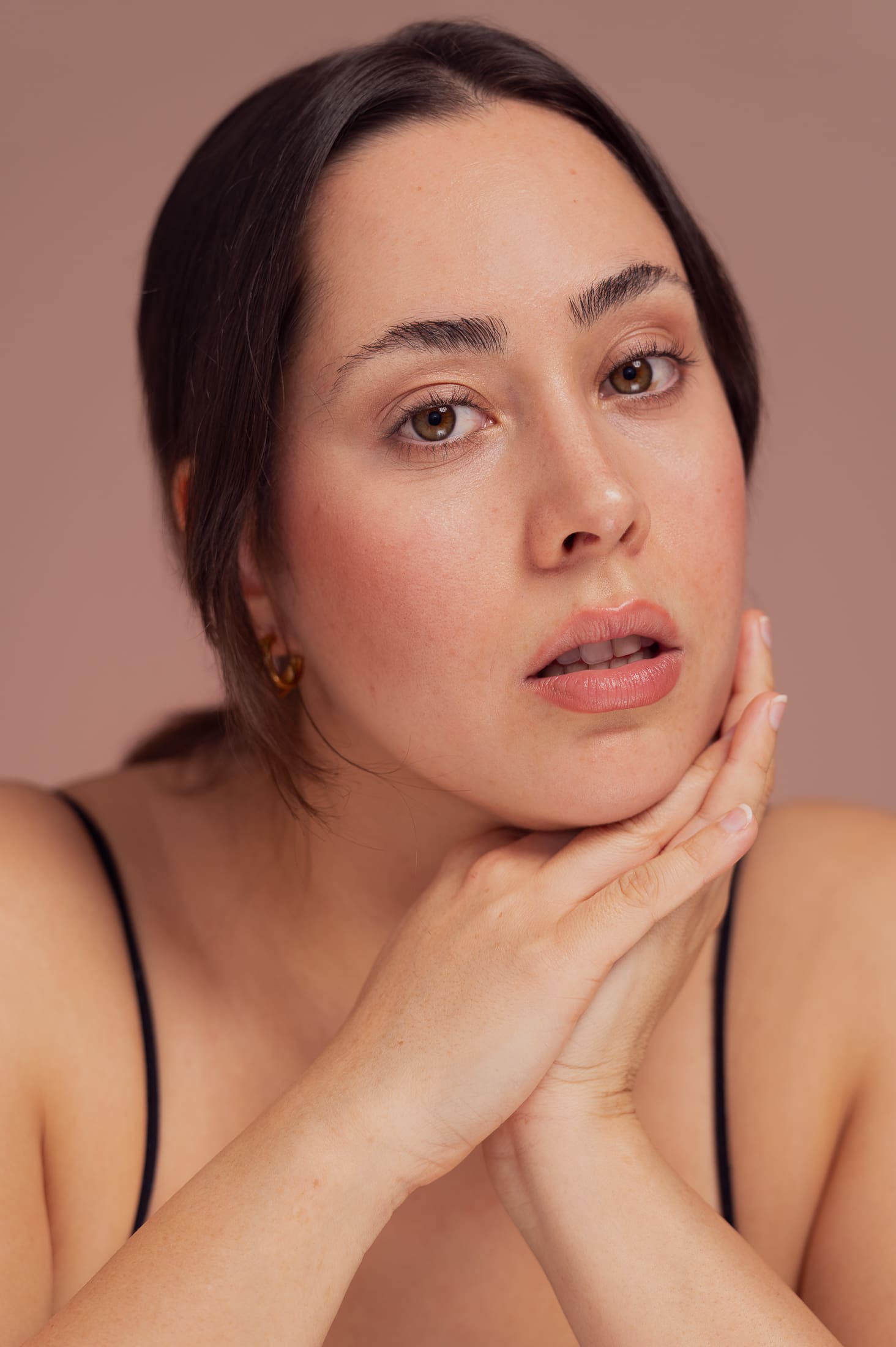Woman leans her chin on her hands with a soft expression, looking to camera in a beauty portrait.