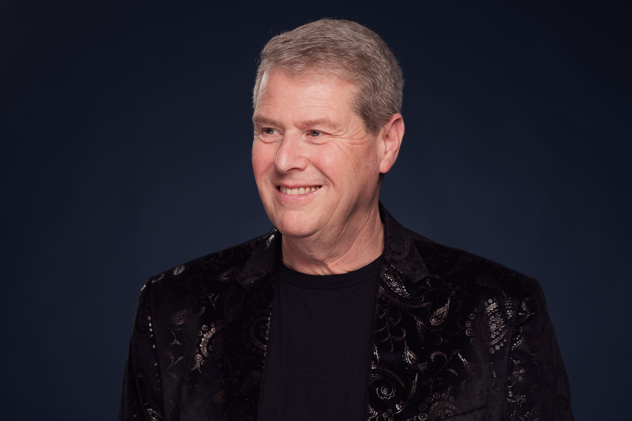 Man smiles away from camera wearing a dark patterned suit jacket, on a navy background.