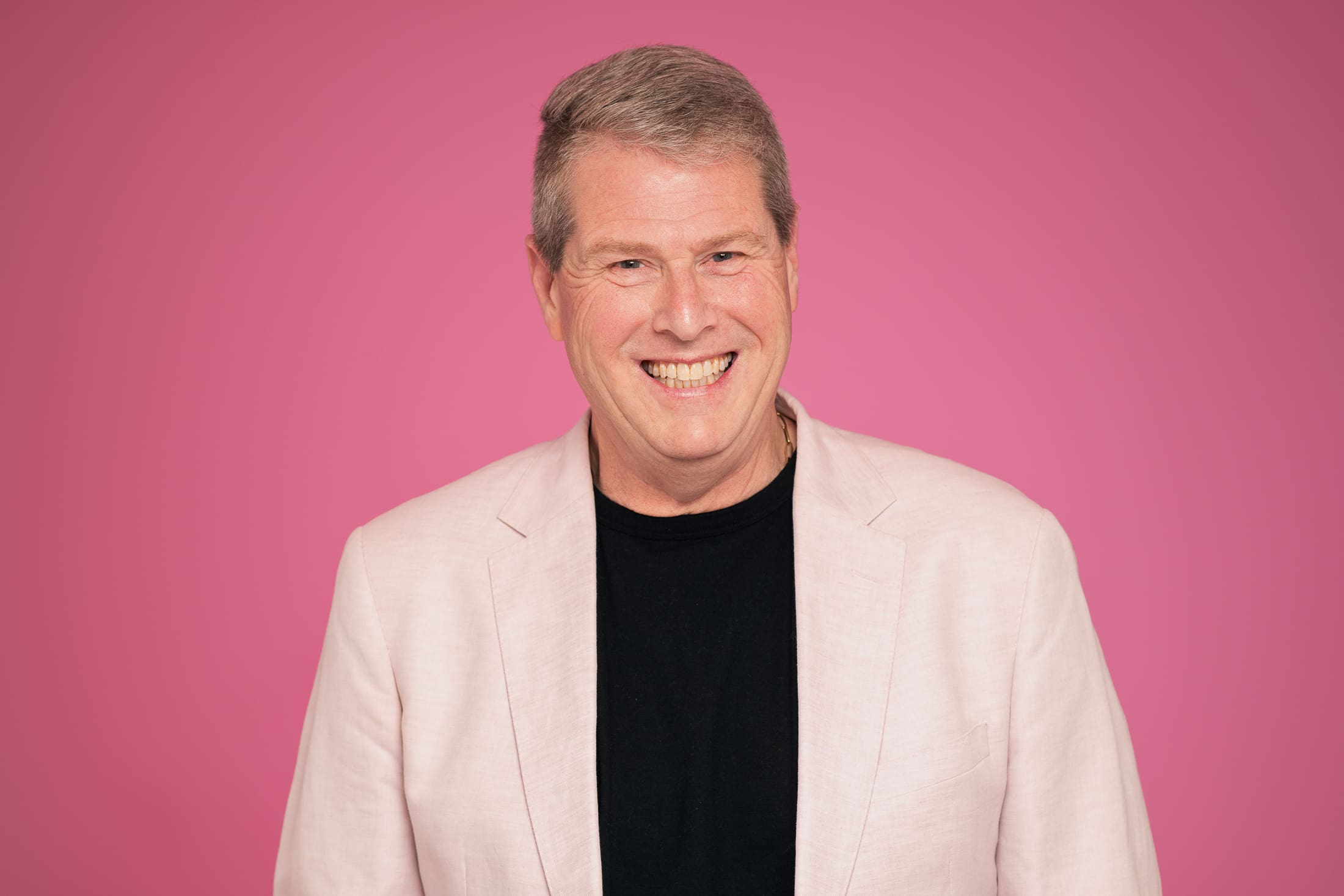 A man wears a light pink blazer and smiles broadly to camera, on a bright pink background.