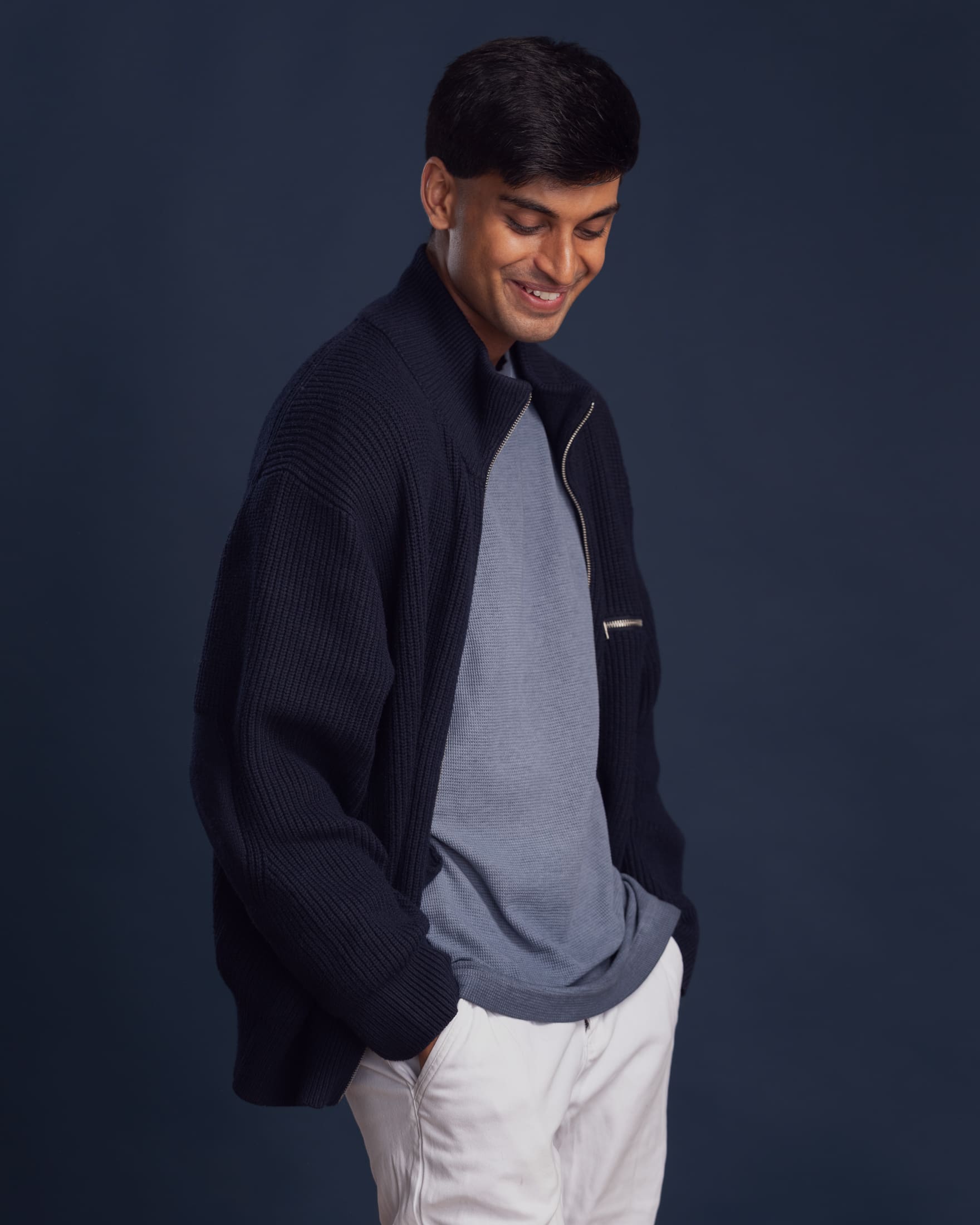 Man laughs away from camera. He stands with his hands in his pockets, wears a navy jacket over a blue tshirt.