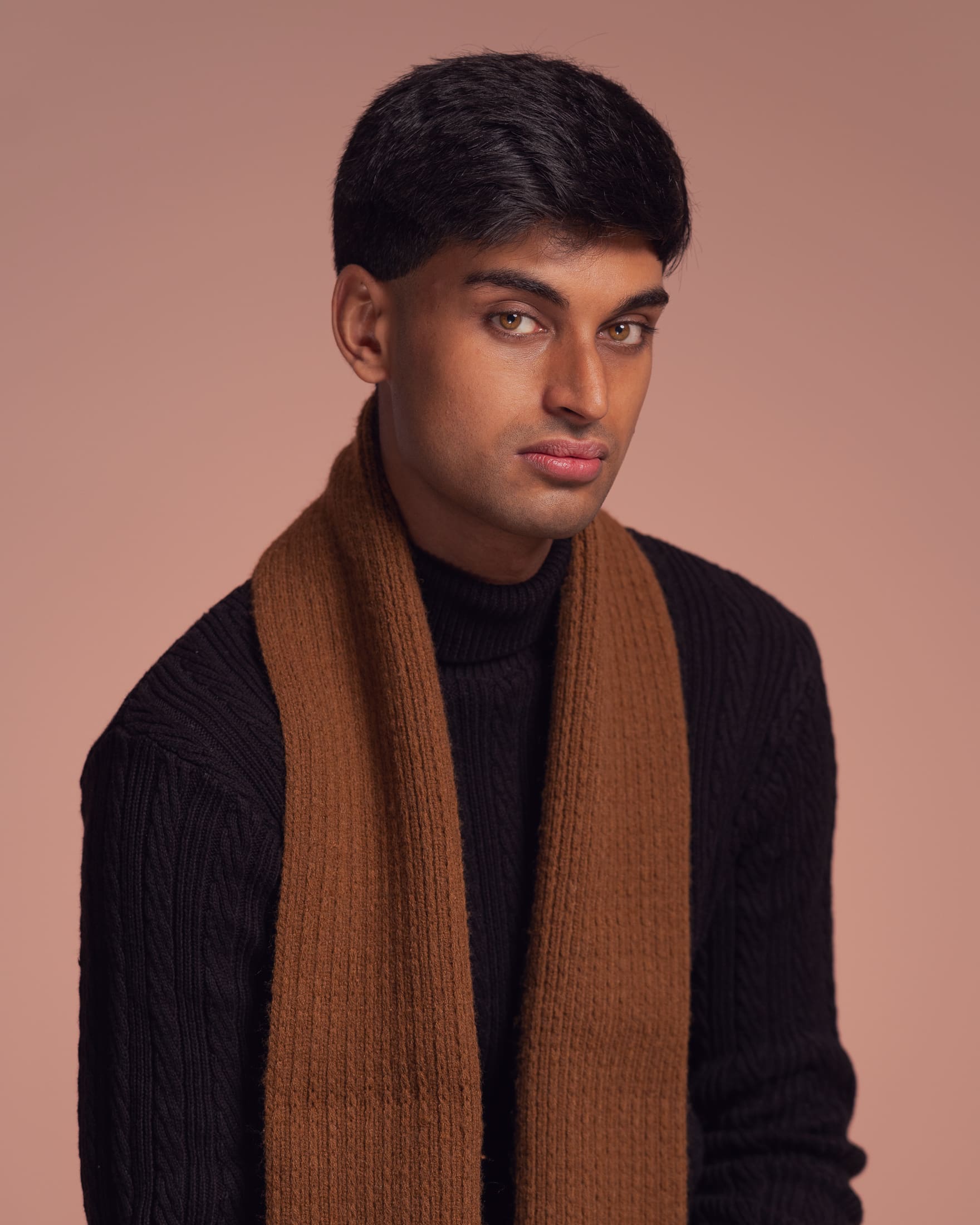 Man looks to camera with a soft but serious expression. He has a brown scarf and is photographed on a soft brown background.