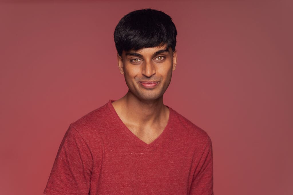 A man smiles to camera. He wears a red v-neck tshirt and is photographed on a muted red background.