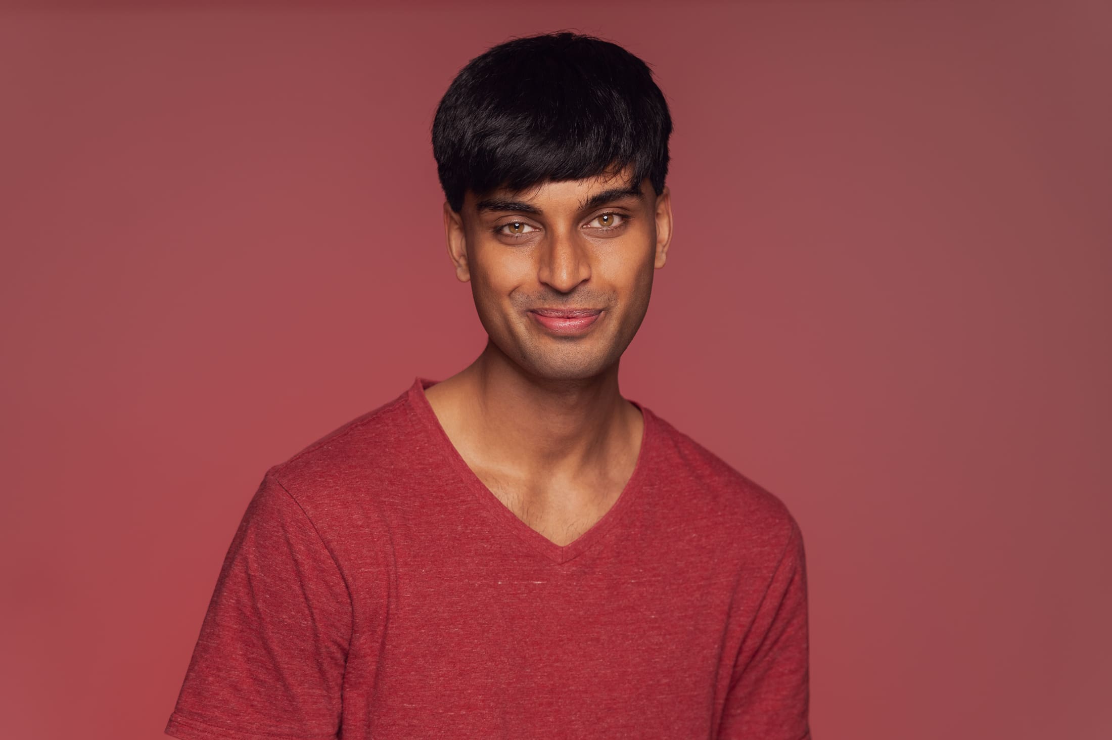 A man smiles to camera. He wears a red v-neck tshirt and is photographed on a muted red background.