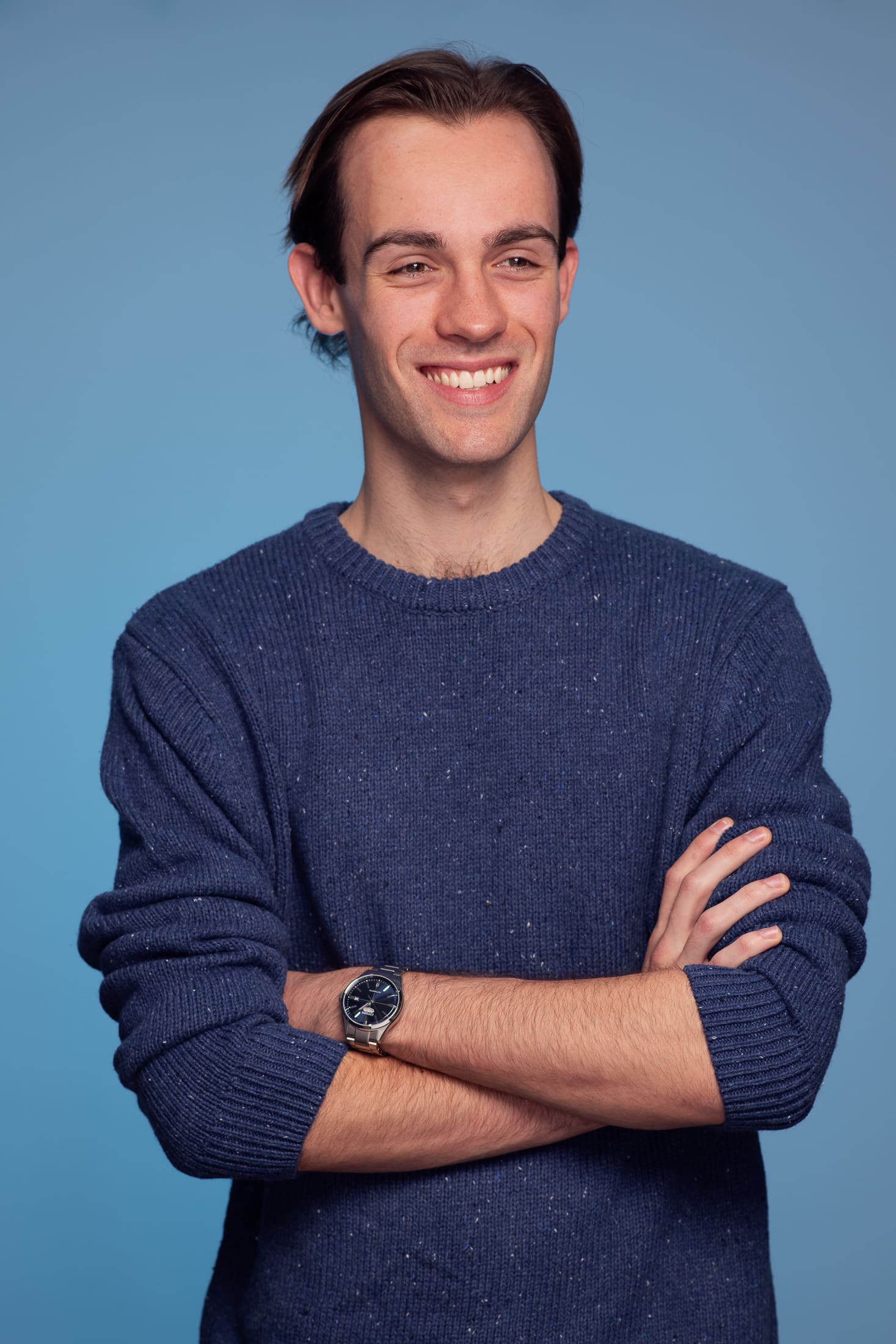 Man laughs away from camera with his arms crossed. he wears a dark blue jumper on a light blue background.