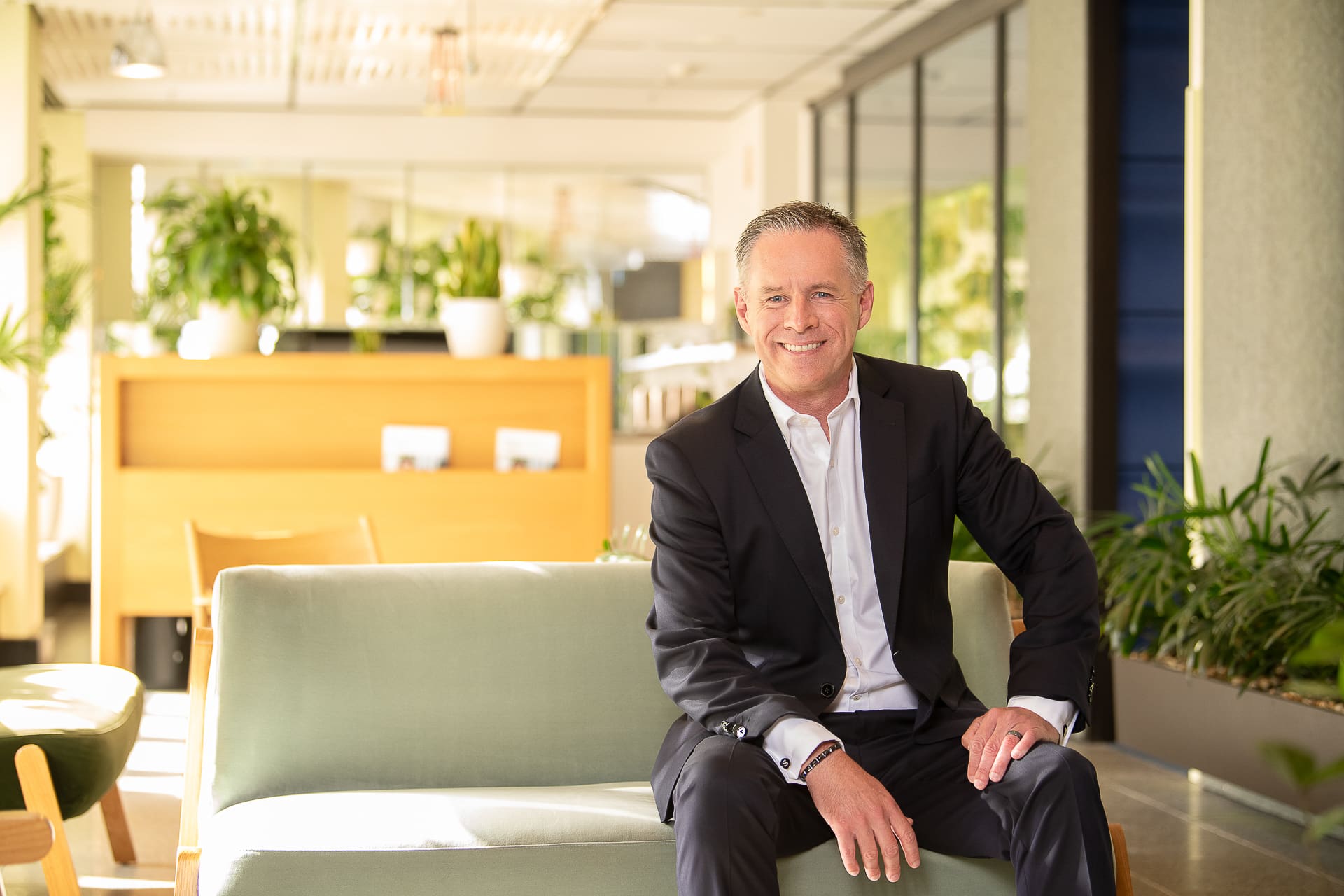 CEO portrait of a man sitting in bright, vibrant office. He has a relaxed pose and smiles to camera.