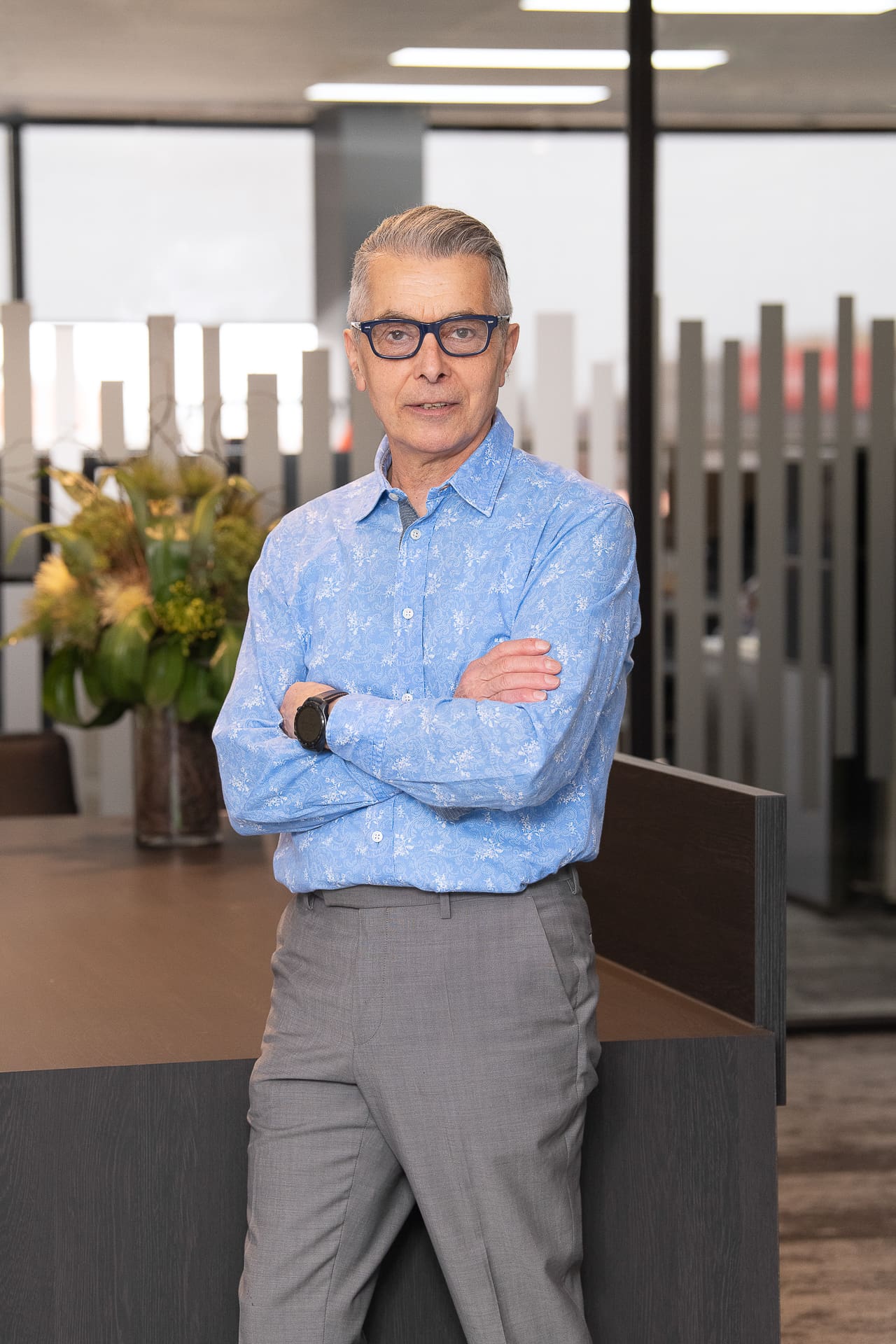 Board executive portrait of man in office setting wearing a sky-blue shirt and dark rimmed glasses. His arms are crossed and he has a serious expression.