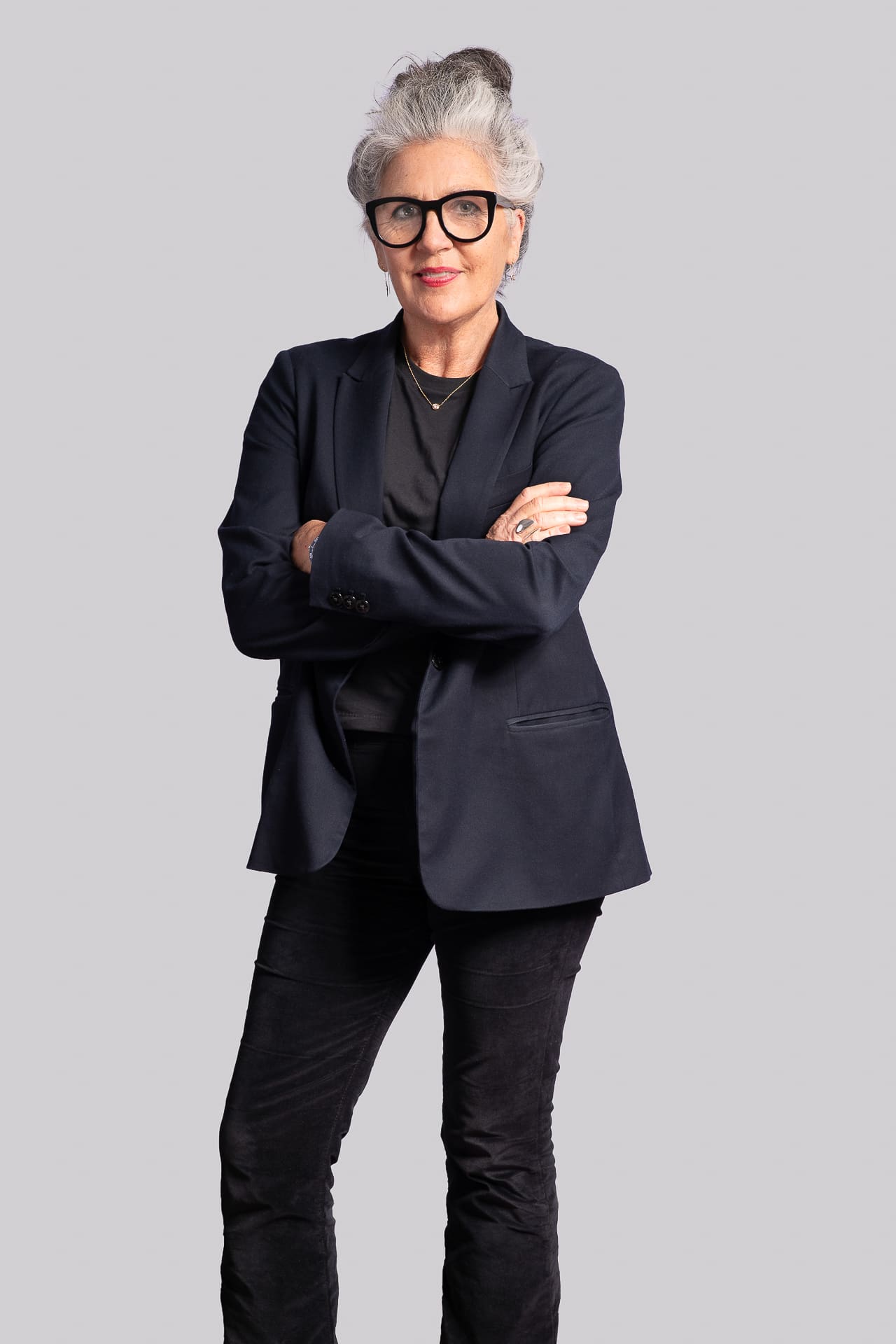 Executive portrait of a woman standing in assertive arms crossed pose. She wears an all-black outfit with tailored blazer and dark rimmed glasses, with an approachable soft smile.