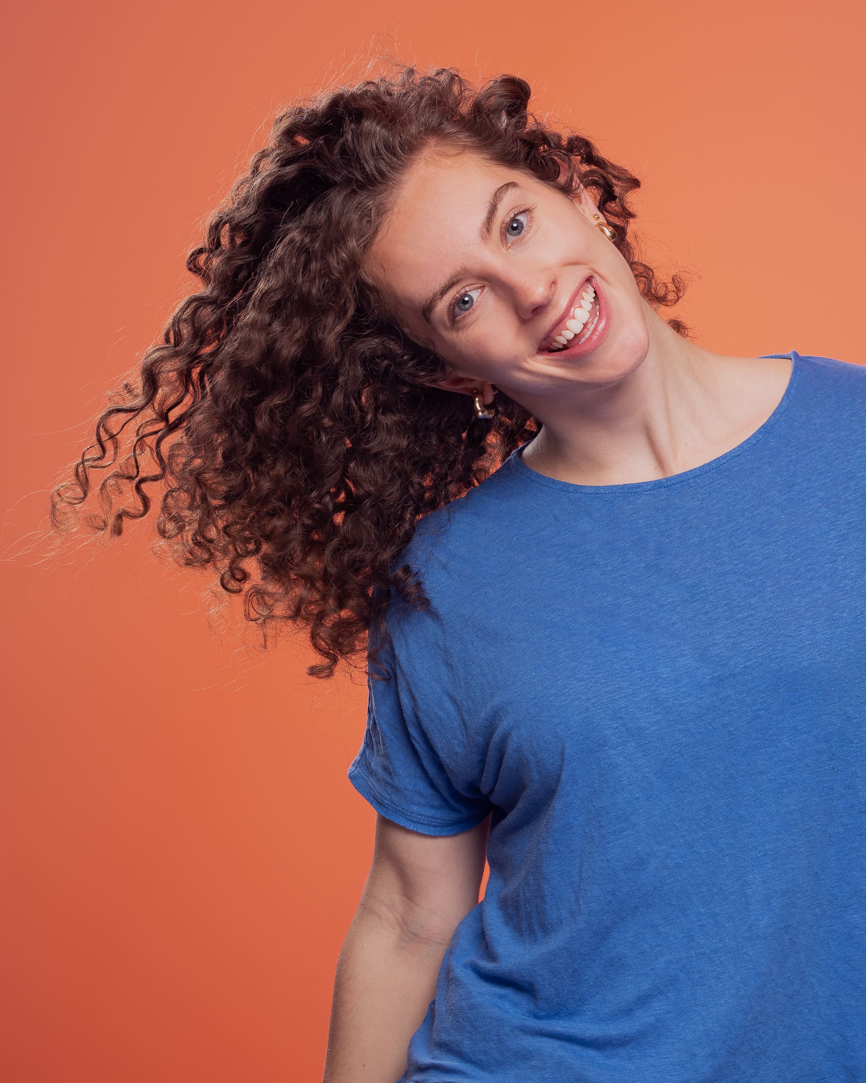 Portrait of a woman smiling to camera. She leans to the left, allowing her curly hair to swing down and away with movement.
