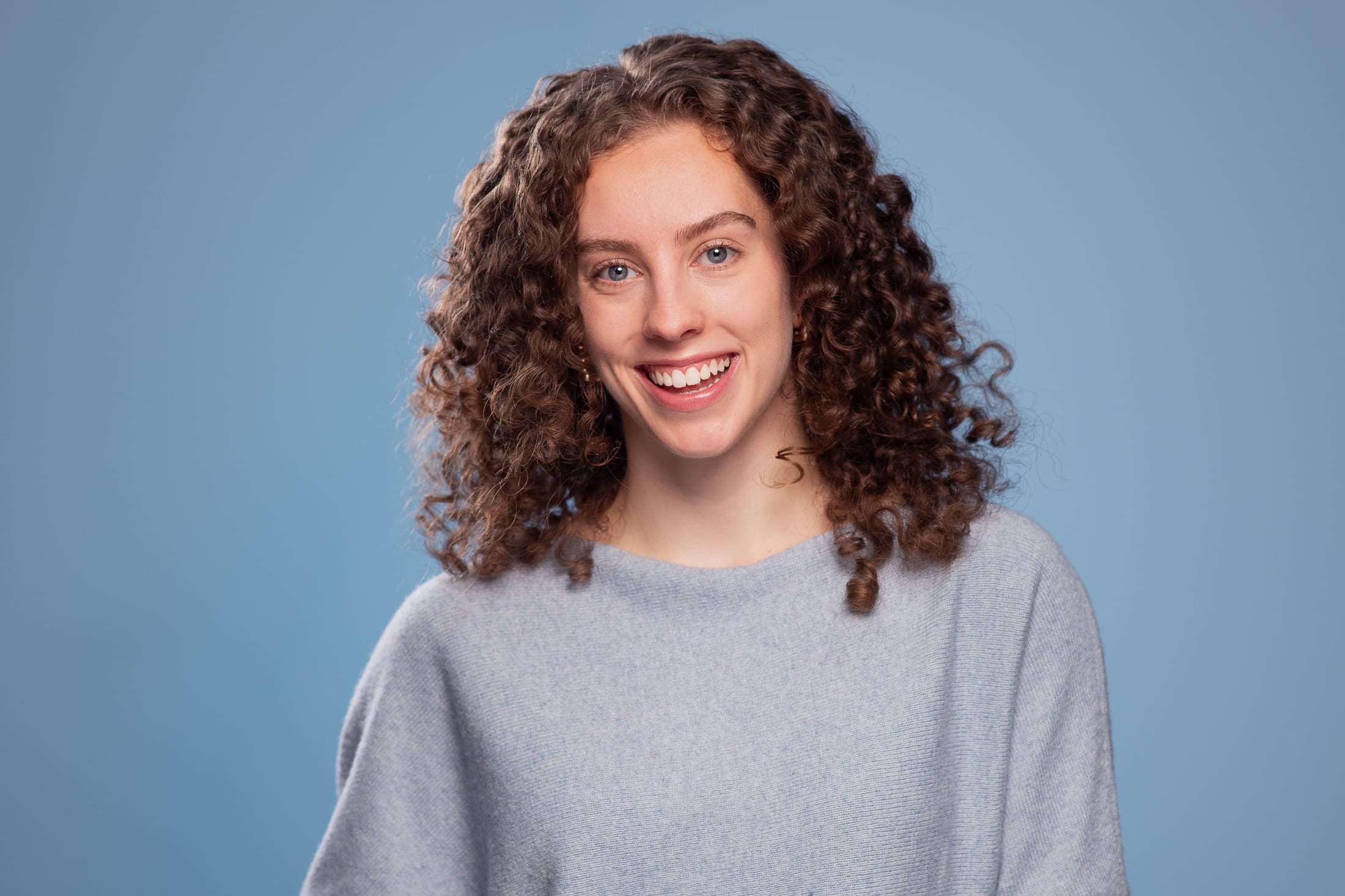 Woman with curly hair smiles widely to camera. She has blue eyes, wears a blue jumper and is on a blue background.