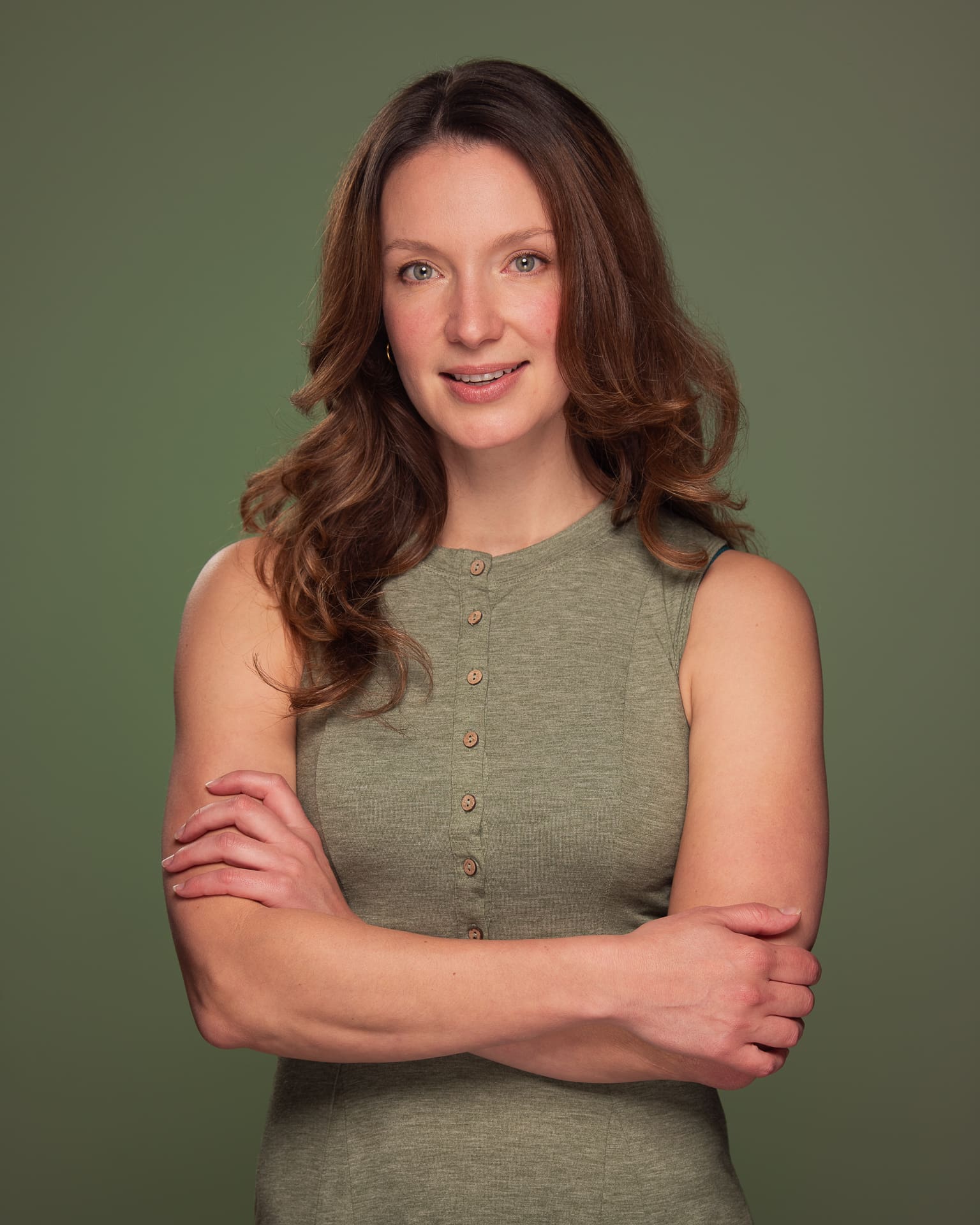 Woman smiles to camera with a relaxed arms crossed pose, wearing a green dress and green background.